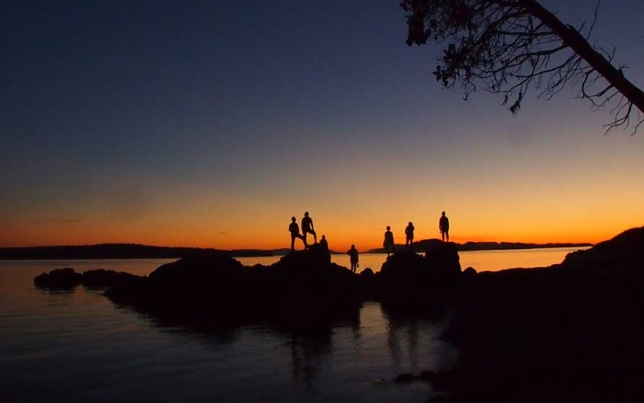 the silhouette of a group of people standing on rocks jutting out of a body of water is illuminated against the sunset.